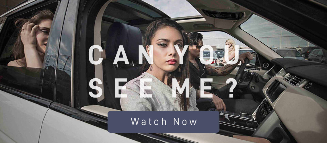 A young woman with makeup on in a grey shirt sits in the front seat of a white SUV while looking at the camera. A man with tattoos is driving the vehicle through a parking lot.